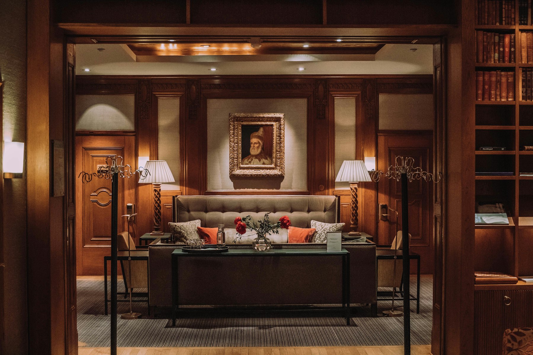A wood-panelled hotel lounge at night, two shaded table lamps and a picture light warming a buttoned sofa below a framed portrait