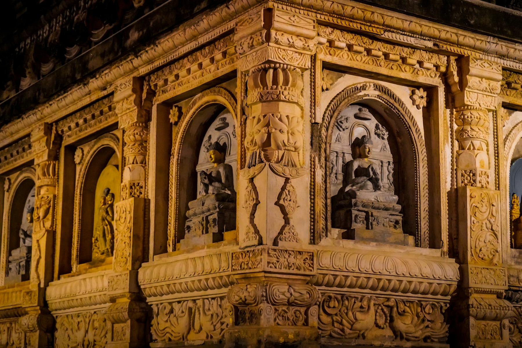 The carved corner of a stone temple at night, warm uplight raking across seated figures set in arched niches