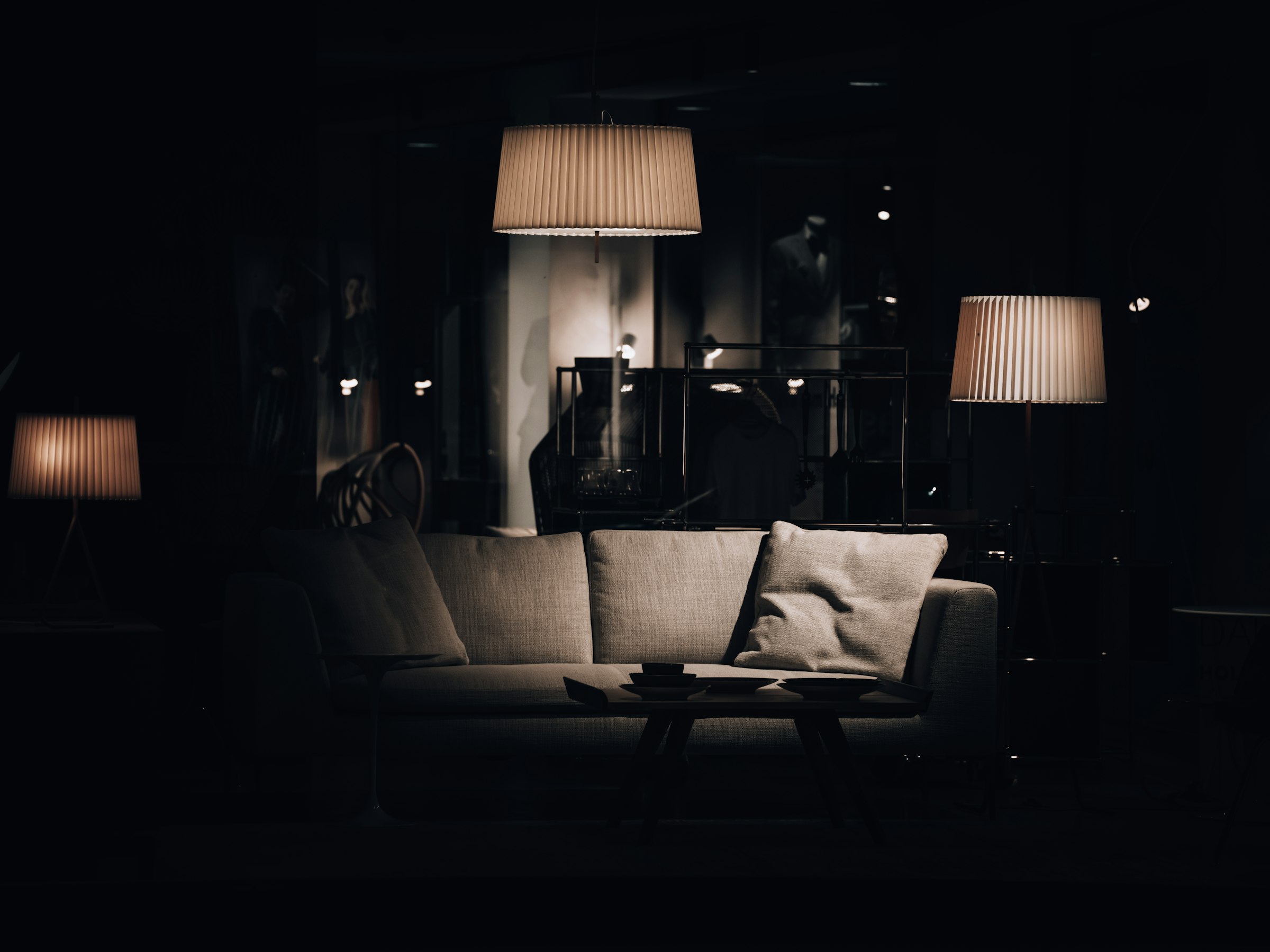 A darkened showroom interior where a pleated pendant and two floor lamps cast warm pools of light over a pale sofa